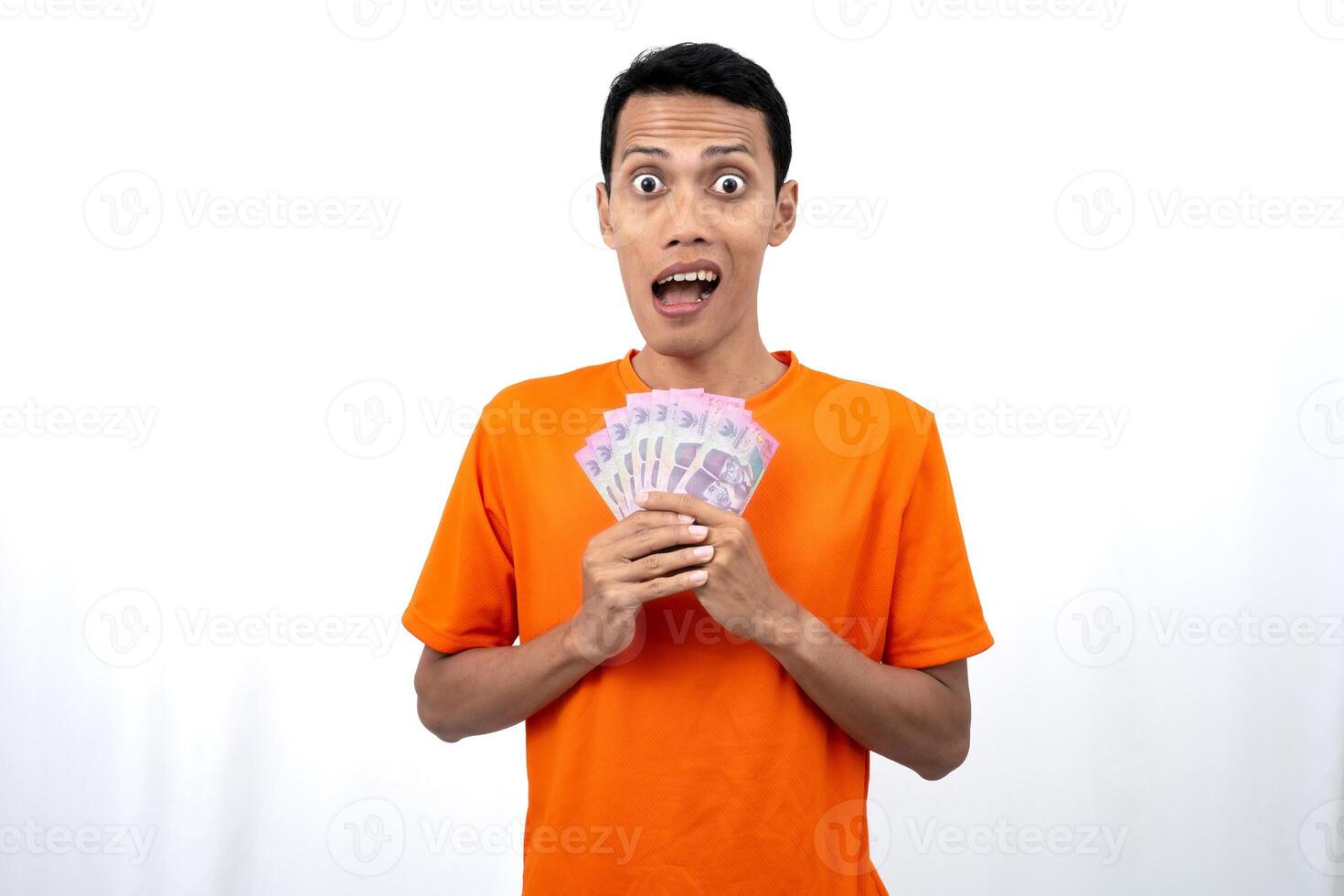 Portrait of a young Asian man wearing an orange t-shirt with a happy, surprised, pleased expression while carrying hundreds of thousands of rupiah. isolated on a white background. photo