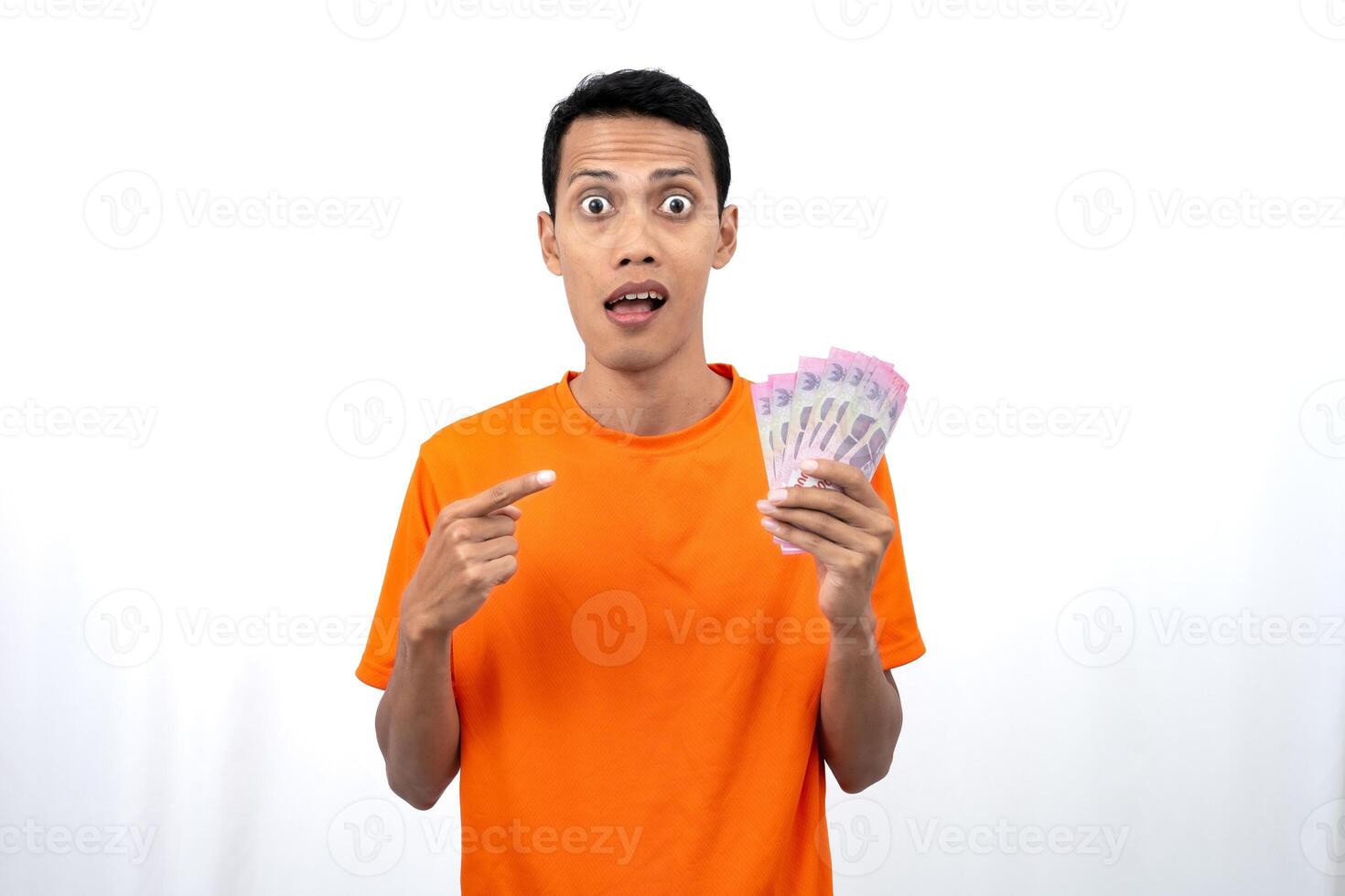 Portrait of a young Asian man wearing an orange t-shirt with a happy, surprised, pleased expression while carrying hundreds of thousands of rupiah. isolated on a white background. photo