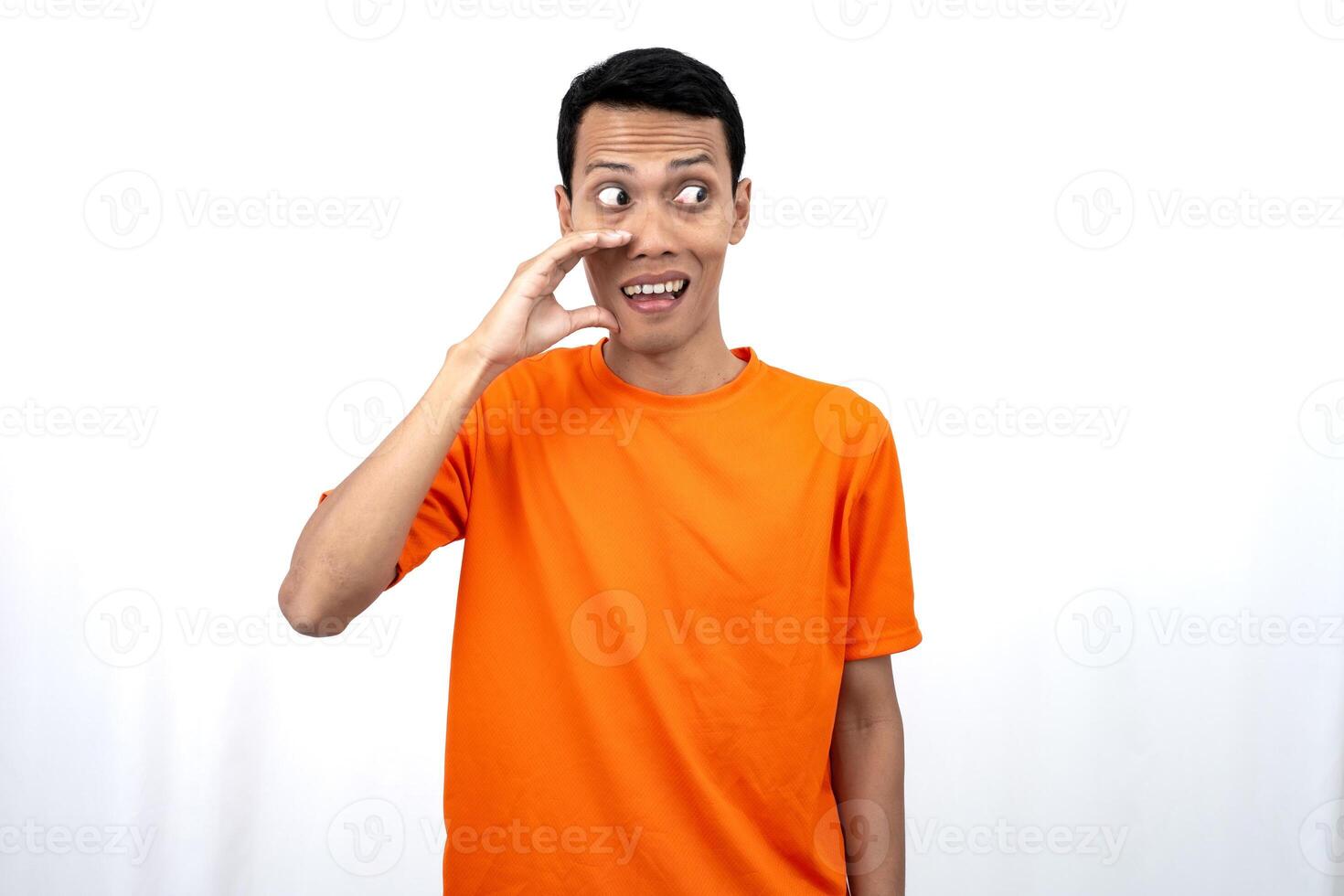 Portrait of a young Asian man wearing an orange t-shirt with his hand near his mouth whispering gossip and telling secrets while sharing news isolated on white background. photo