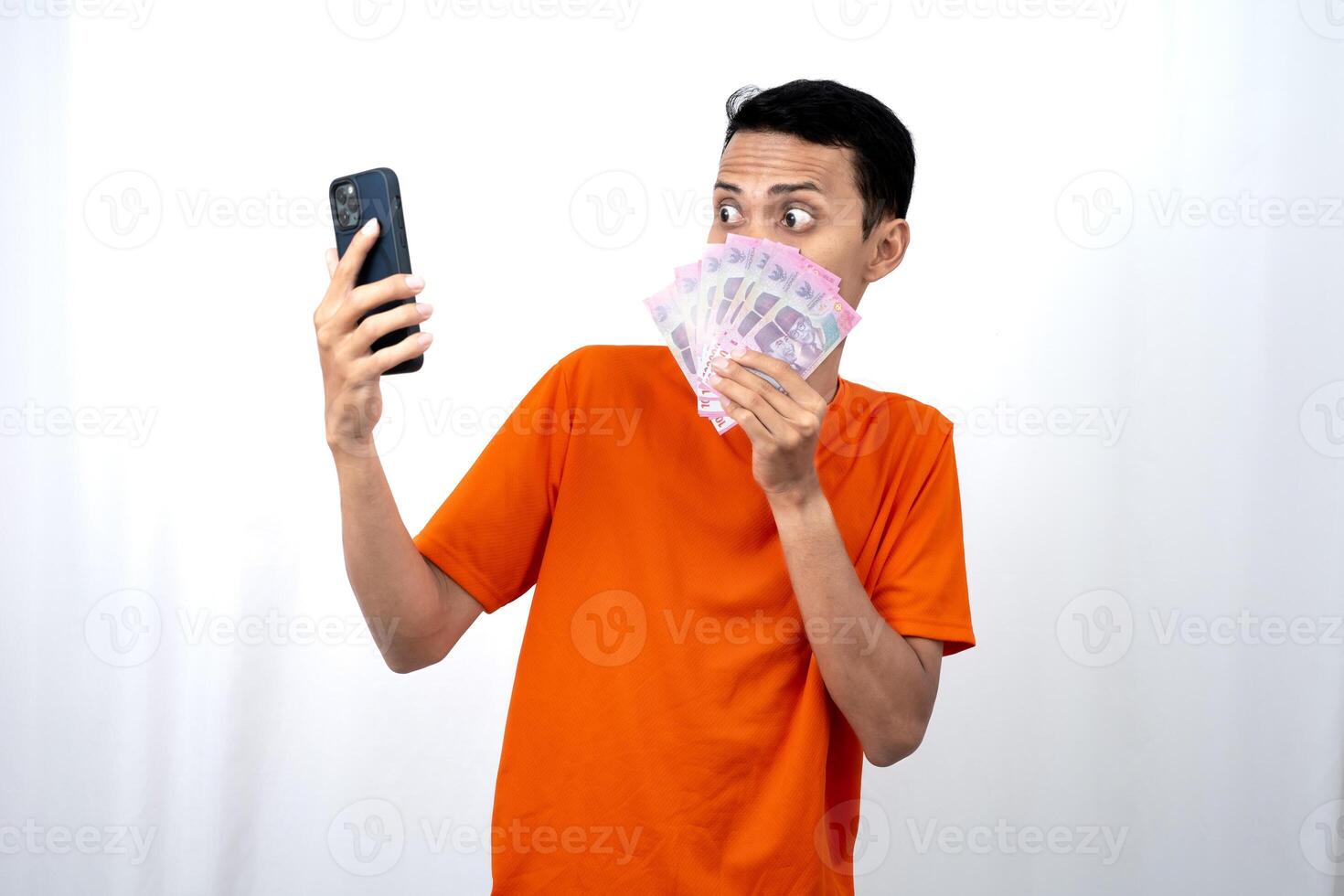 Portrait of a young Asian man wearing an orange t-shirt with a happy, surprised, pleased expression while carrying a smartphone and hundreds of thousands of rupiah. isolated on a white background. photo
