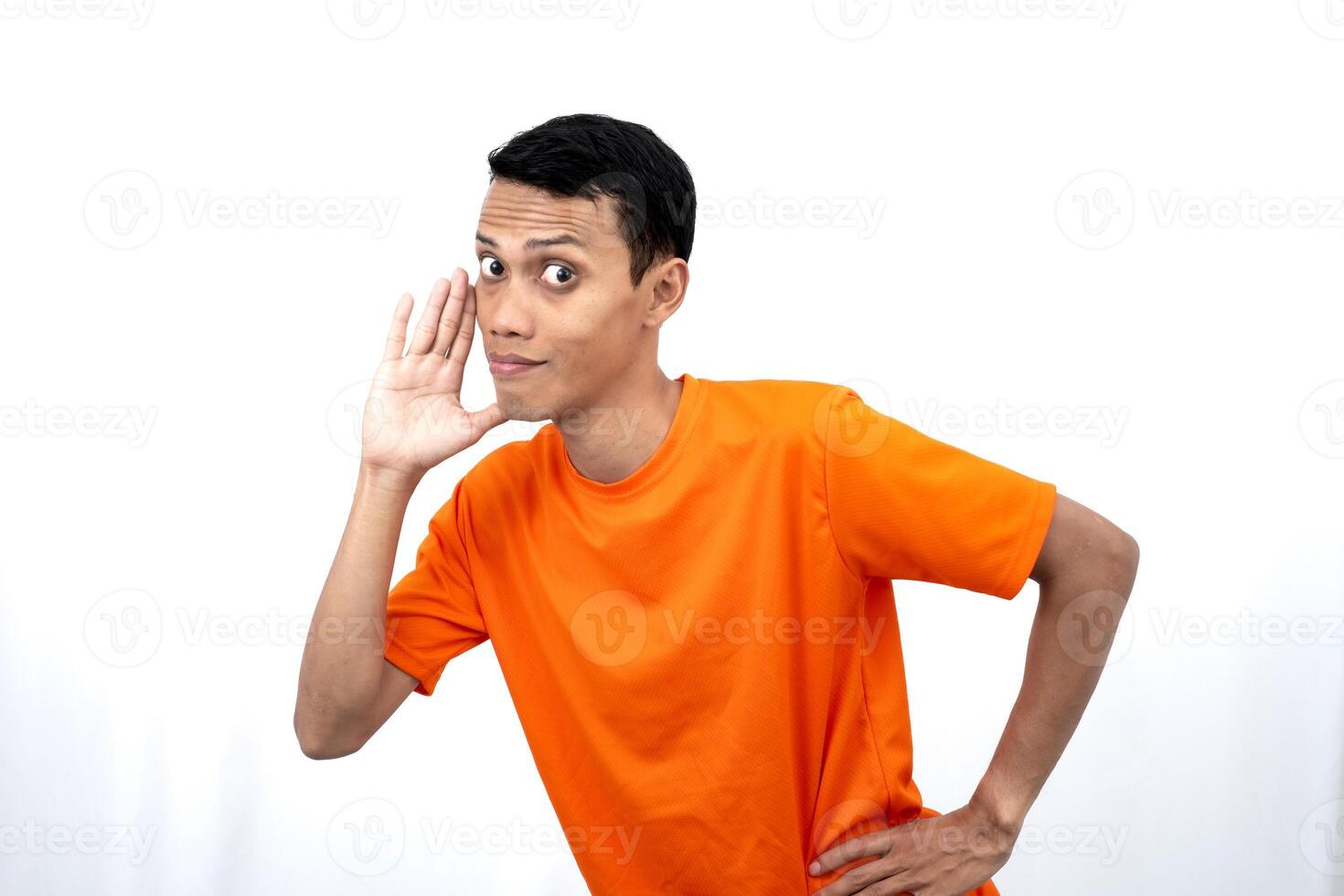 Portrait of a young Asian man wearing an orange t-shirt with his hand near his mouth whispering gossip and telling secrets while sharing news isolated on white background. photo