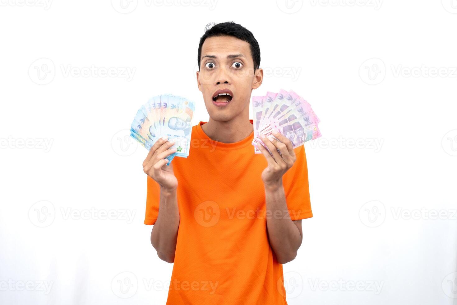 Portrait of a young Asian man wearing an orange t-shirt with a happy, surprised, pleased expression while carrying hundreds of thousands of rupiah. isolated on a white background. photo