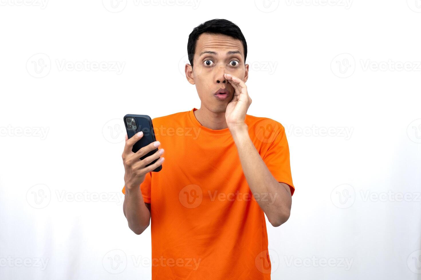 Portrait of a young Asian man wearing an orange t-shirt with his hand near his mouth whispering gossip and telling secrets while holding a smartphone. Sharing news isolated on white background. photo
