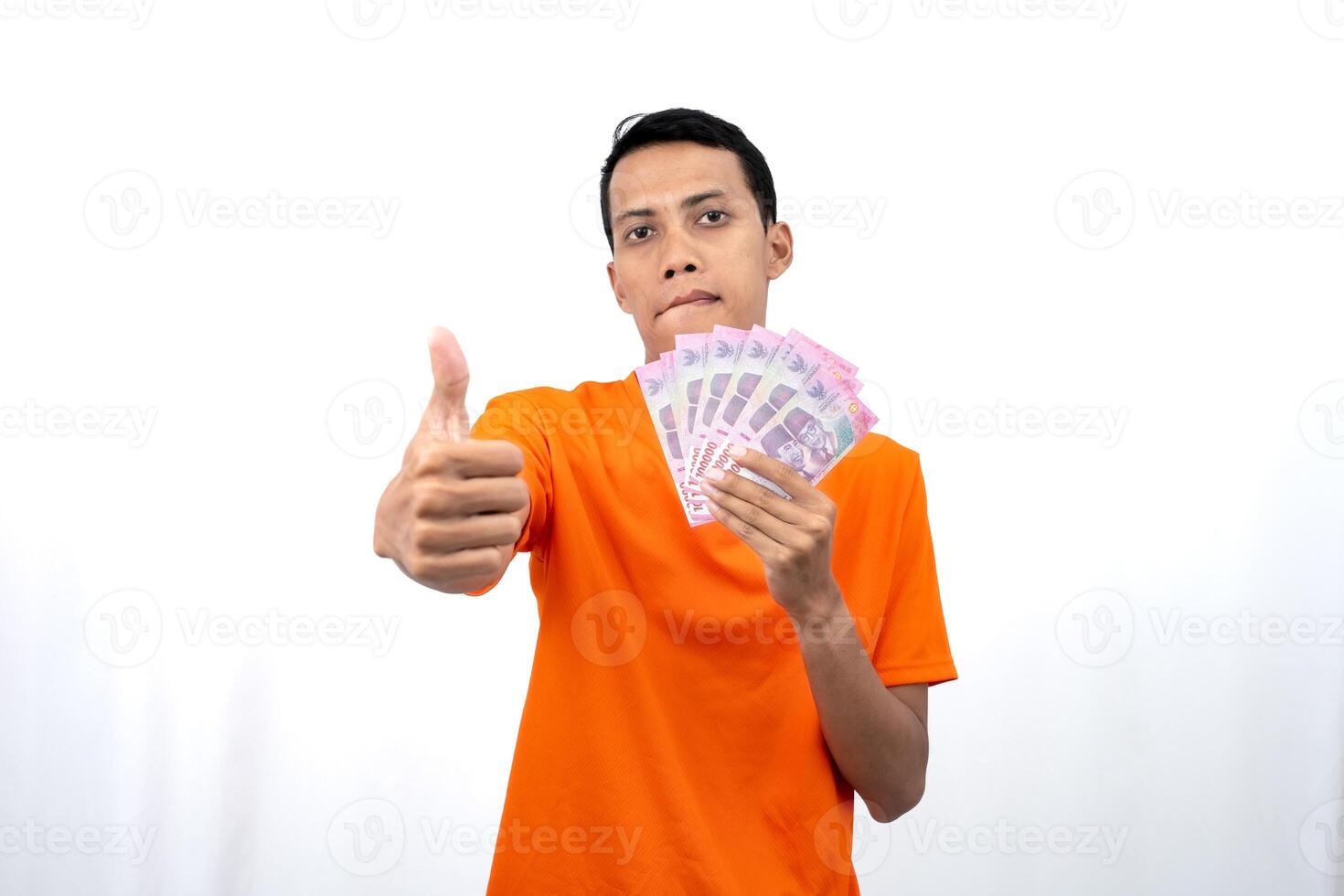 Portrait of a young Asian man wearing an orange t-shirt with a happy, surprised, pleased expression while carrying hundreds of thousands of rupiah. isolated on a white background. photo