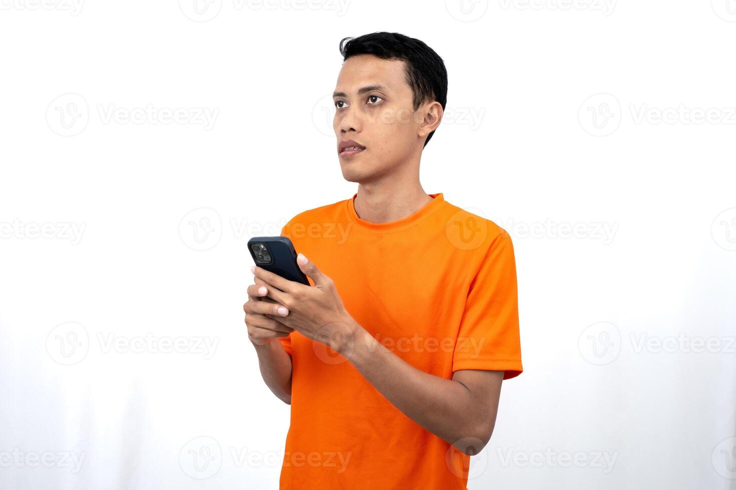 Portrait of a young Asian man wearing an orange t-shirt showing thinking, while holding a smartphone. Isolated on white background. photo