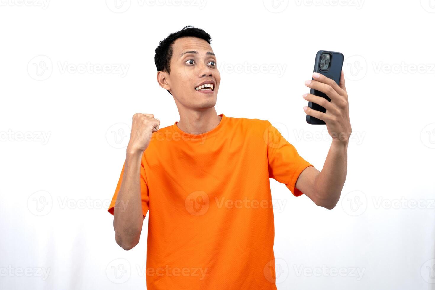 Portrait of a young Asian man wearing an orange t-shirt with a happy expression, surprised to get happy news via smartphone. Isolated on white background. photo