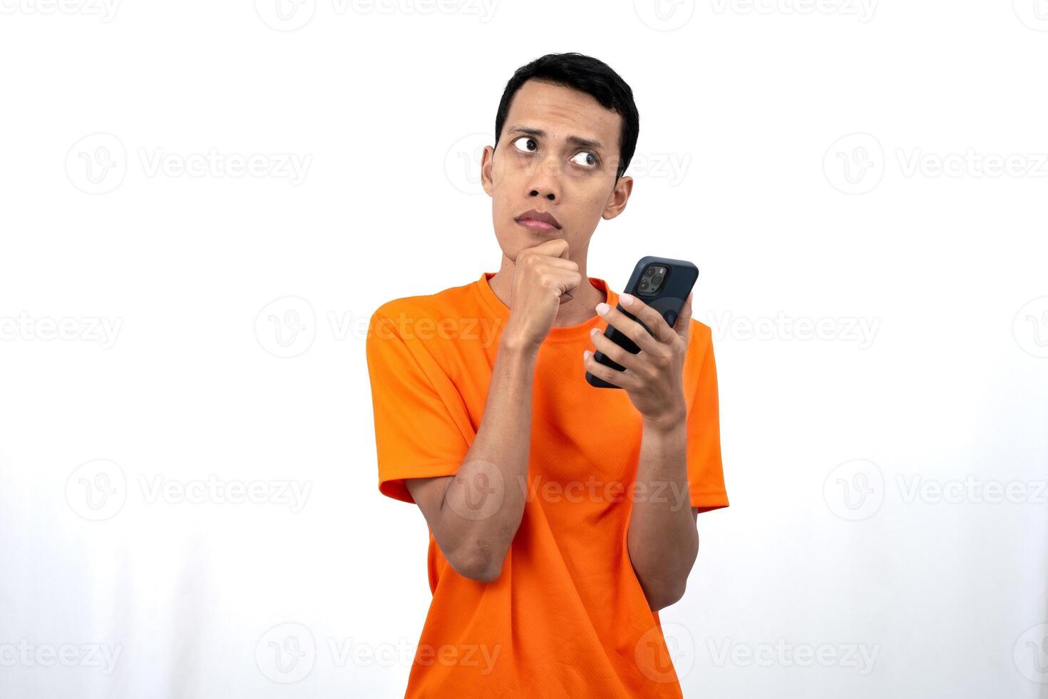 Portrait of a young Asian man wearing an orange t-shirt showing thinking, while holding a smartphone. Isolated on white background. photo