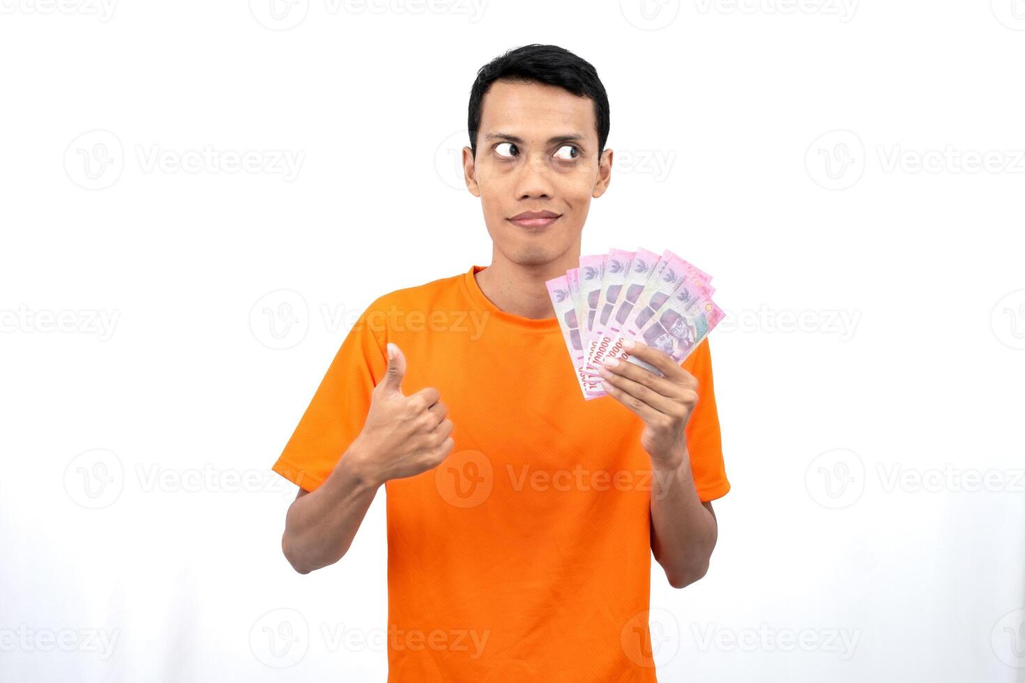 Portrait of a young Asian man wearing an orange t-shirt with a happy, surprised, pleased expression while carrying hundreds of thousands of rupiah. isolated on a white background. photo