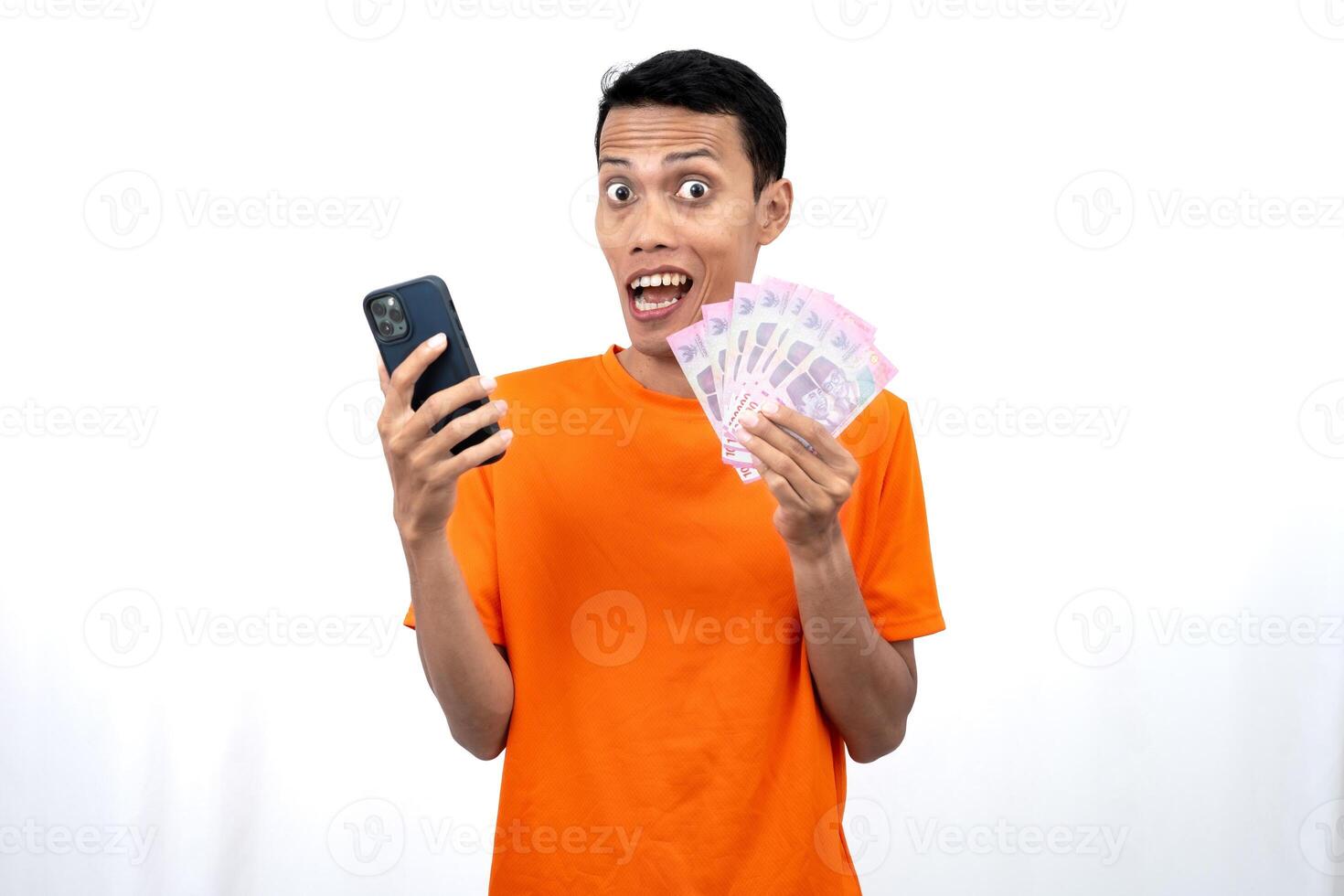 Portrait of a young Asian man wearing an orange t-shirt with a happy, surprised, pleased expression while carrying a smartphone and hundreds of thousands of rupiah. isolated on a white background. photo