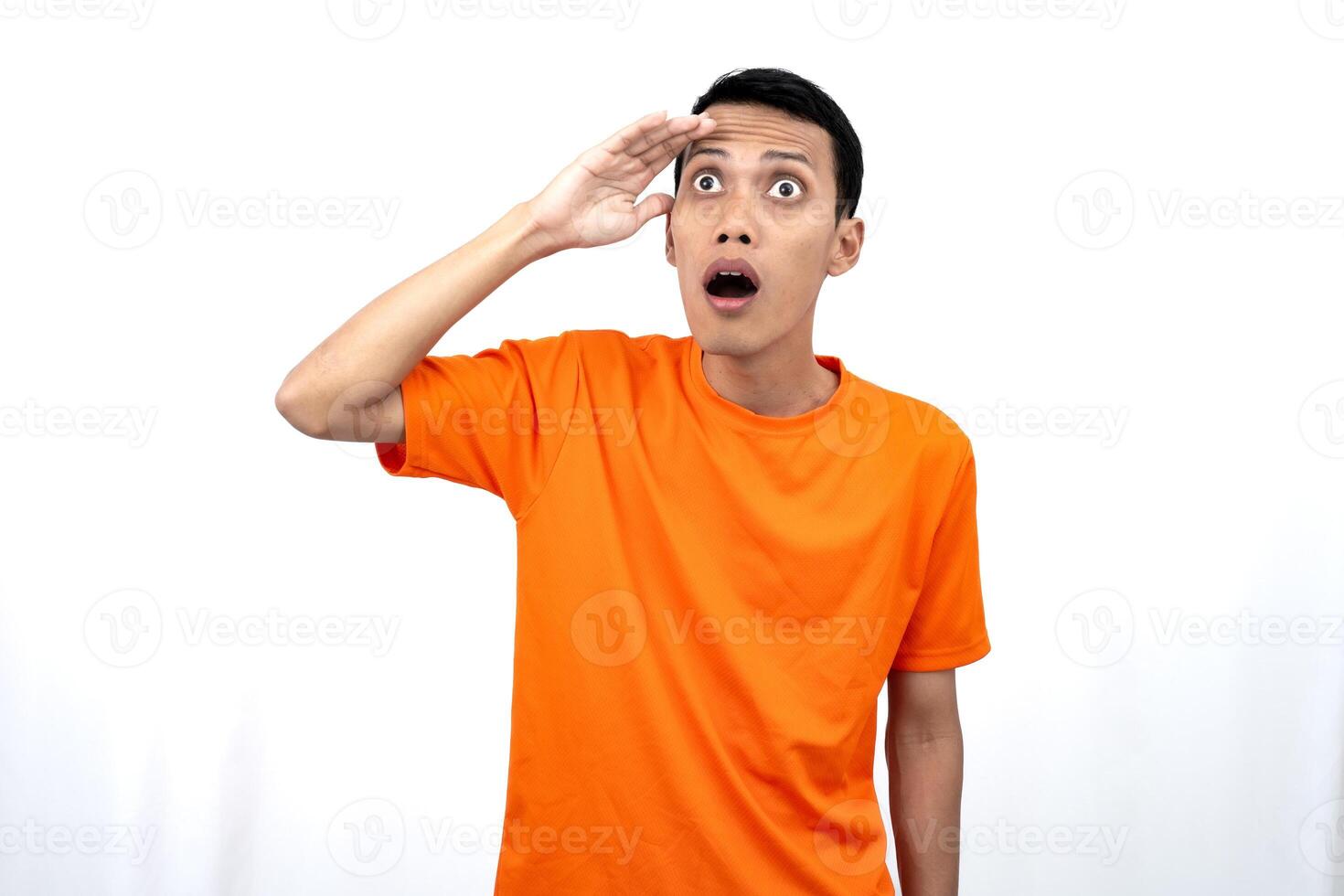 Portrait of a young Asian man wearing an orange t-shirt looking surprised, shocked and shocked. Hands are near eyes, isolated on white background. photo