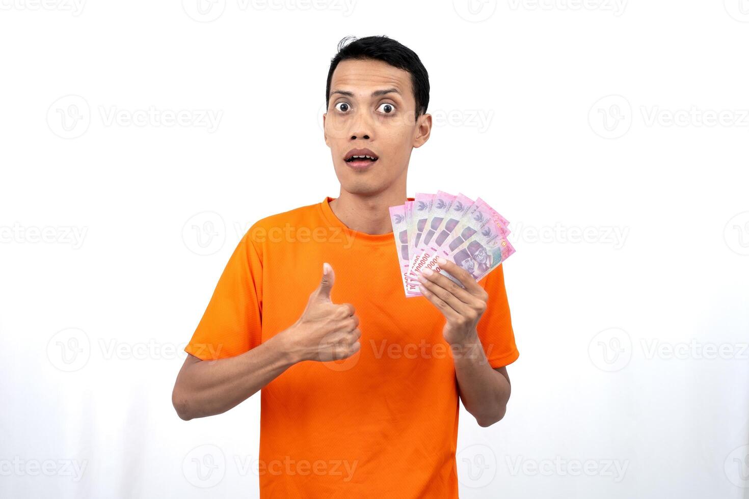 Portrait of a young Asian man wearing an orange t-shirt with a happy, surprised, pleased expression while carrying hundreds of thousands of rupiah. isolated on a white background. photo