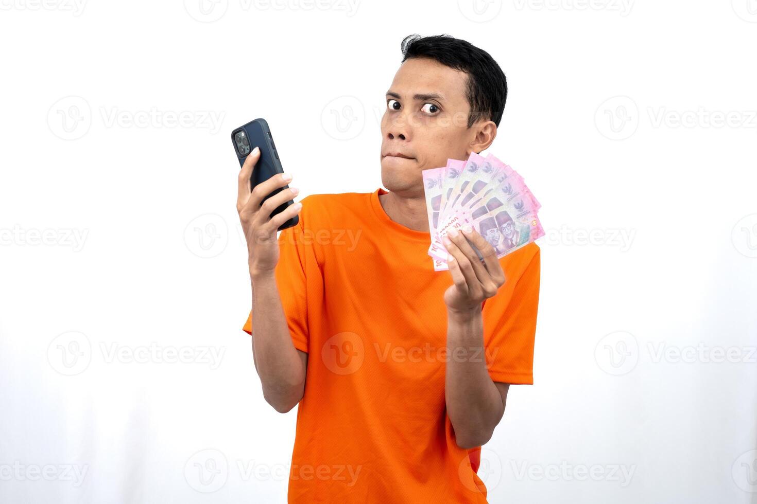 Portrait of a young Asian man wearing an orange t-shirt with a happy, surprised, pleased expression while carrying a smartphone and hundreds of thousands of rupiah. isolated on a white background. photo