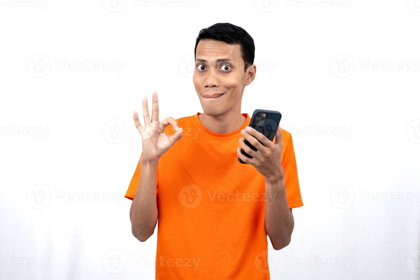 Portrait of a young Asian man wearing an orange t-shirt giving an okay sign, agree, while holding a smartphone. Isolated on white background. photo