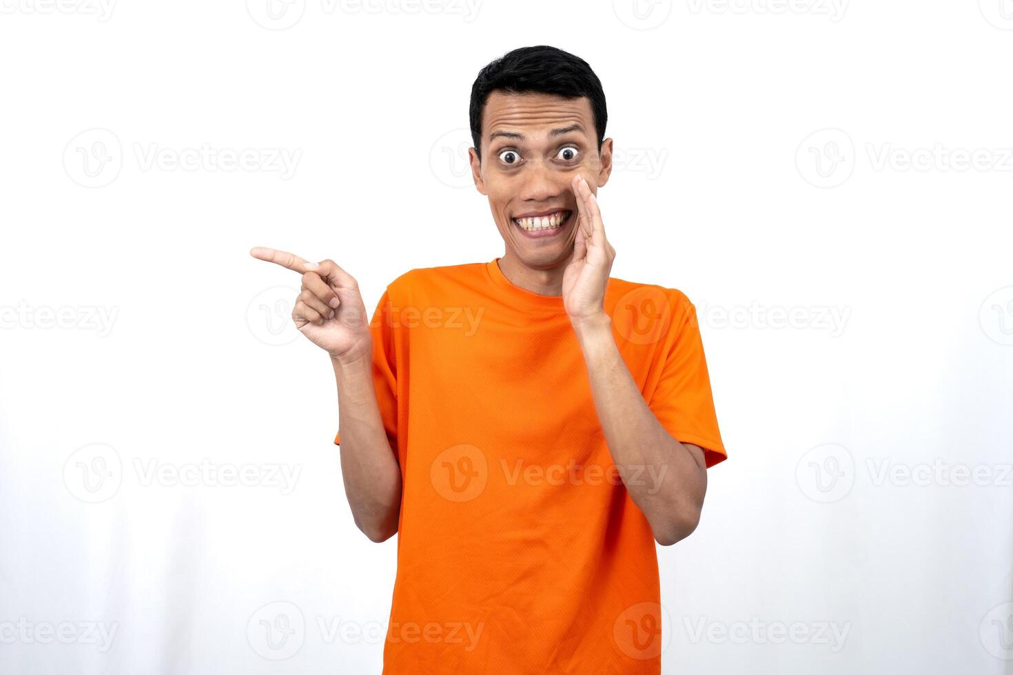 Portrait of a young Asian man wearing an orange t-shirt with his hand near his mouth in a whispering pose while pointing to the side of the copy space. Isolated on white background. photo