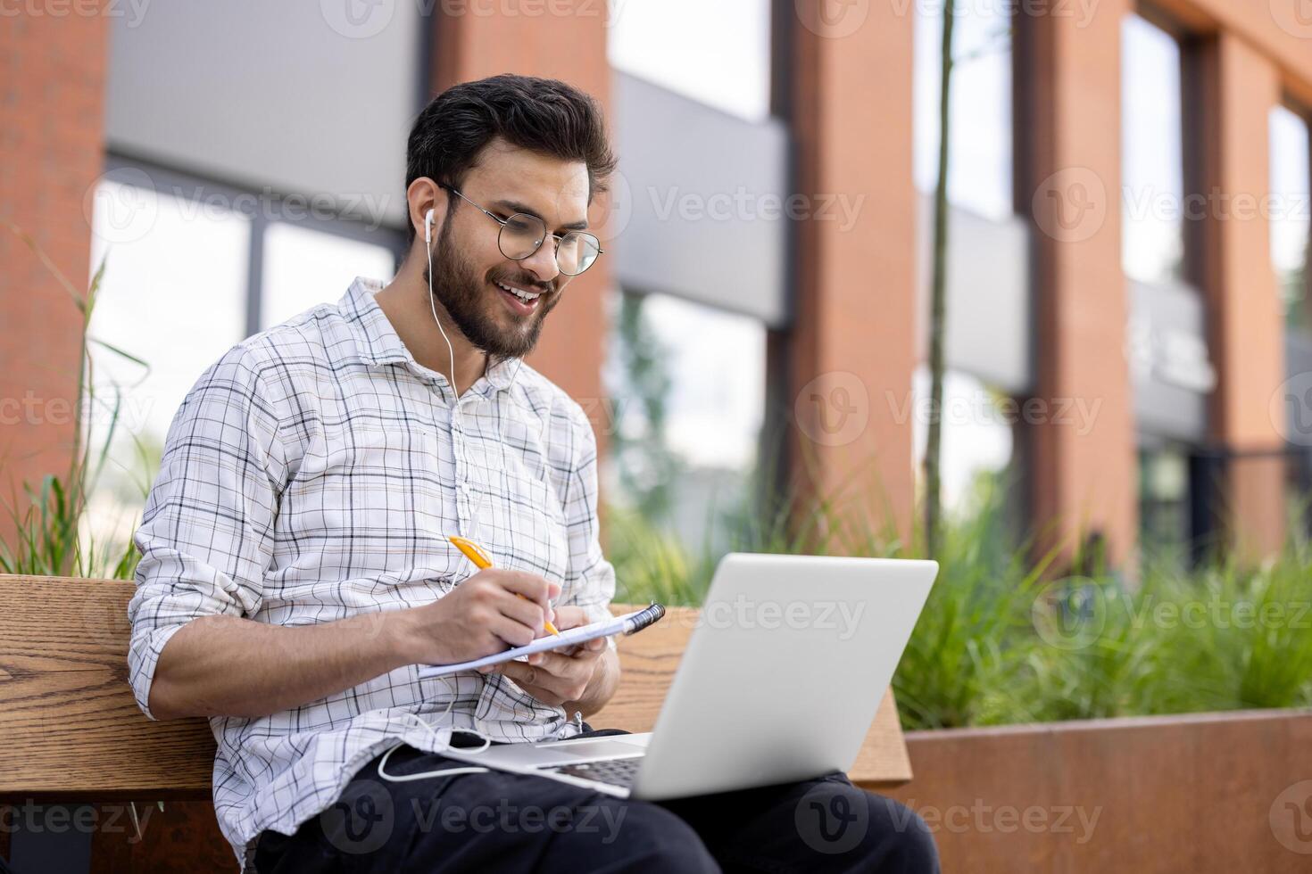 Smiling young Indian man sitting on a bench outside wearing headphones, holding a laptop on his lap, talking on a call, making notes in a notebook. photo