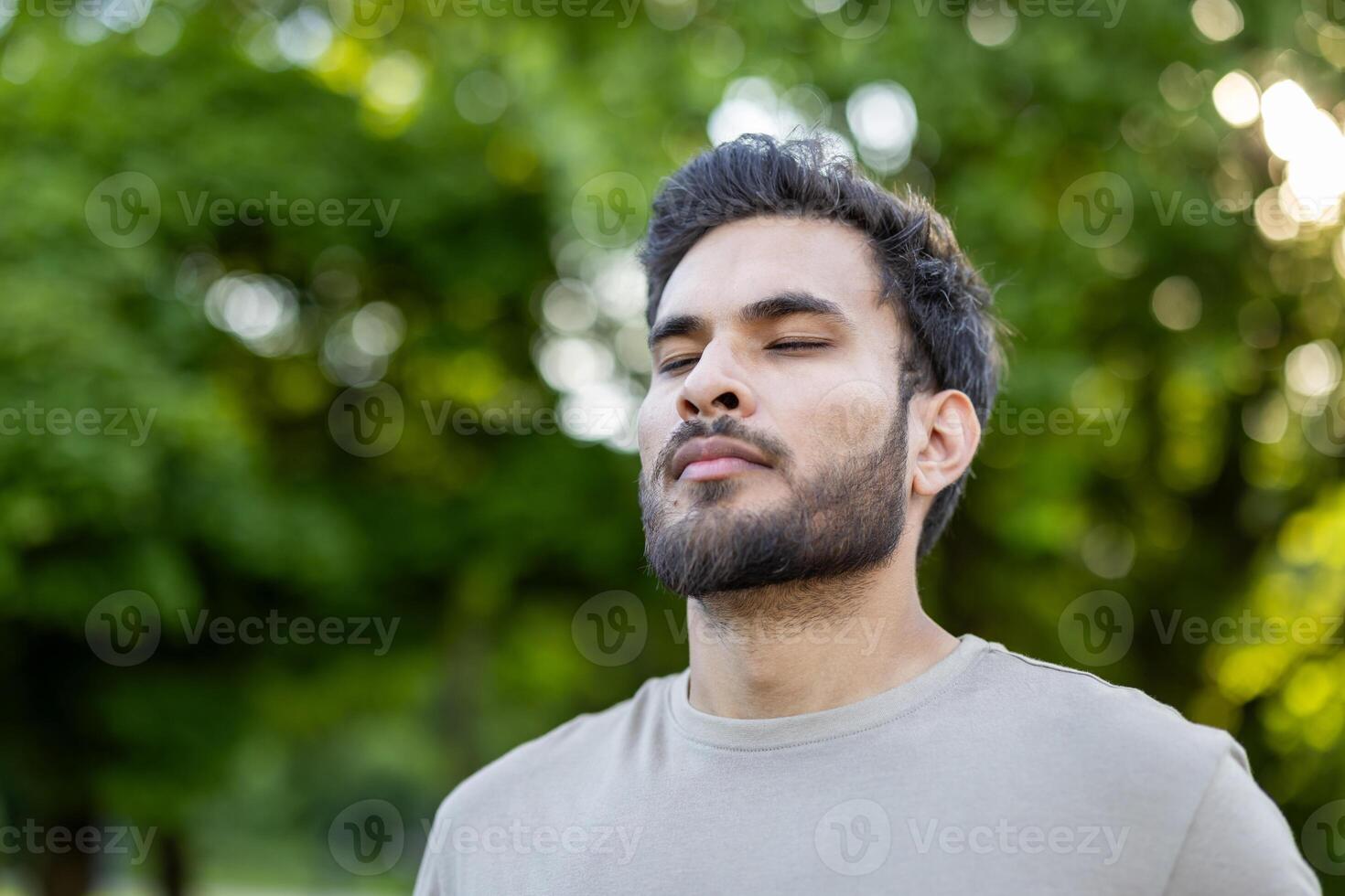 Young man stands with closed eyes embracing tranquility in serene outdoor setting. Surrounded by lush green trees, he reflects calmness and peace in natural environment. photo