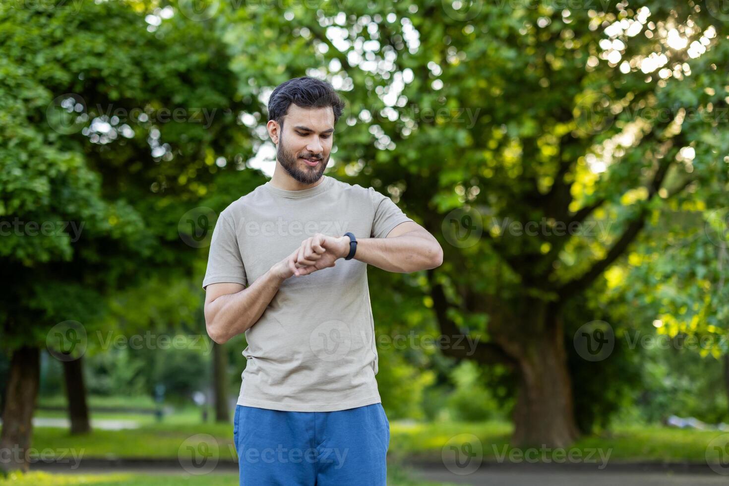 Young man outdoors checking smartwatch during fitness routine. Surrounded by lush greenery, enjoying active lifestyle in park setting. Focus on technology and exercise in natural environment photo