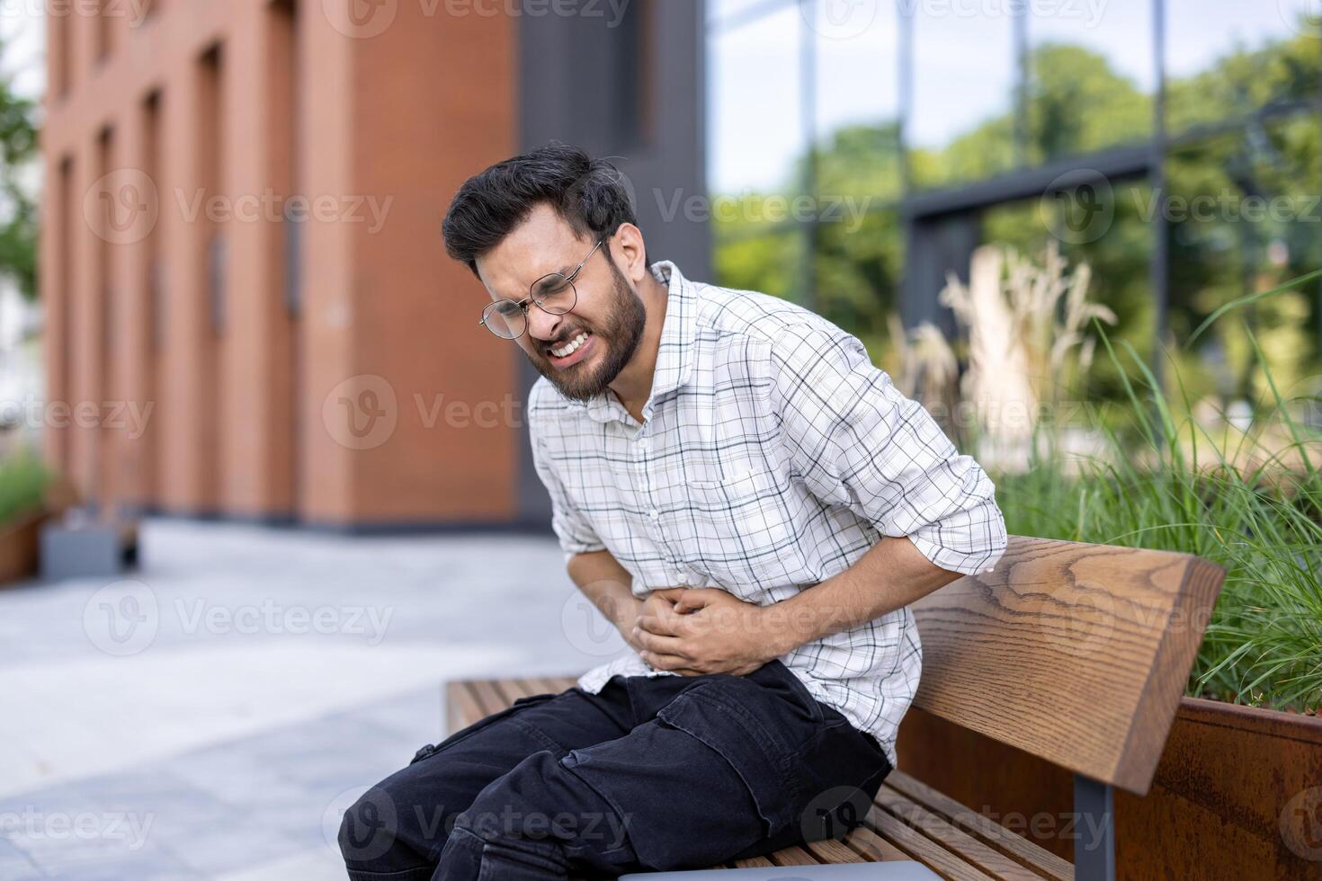 Contorted from severe pain, a young Indian man sits on a bench in the street and holds his stomach with his hands. photo
