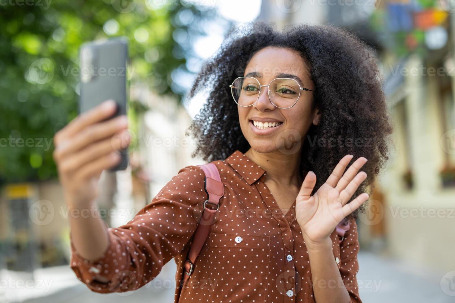 Close-up photo of a young African-American woman wearing glasses and carrying a backpack standing on a city street and talking on a call from her phone, greeting and waving at the camera.