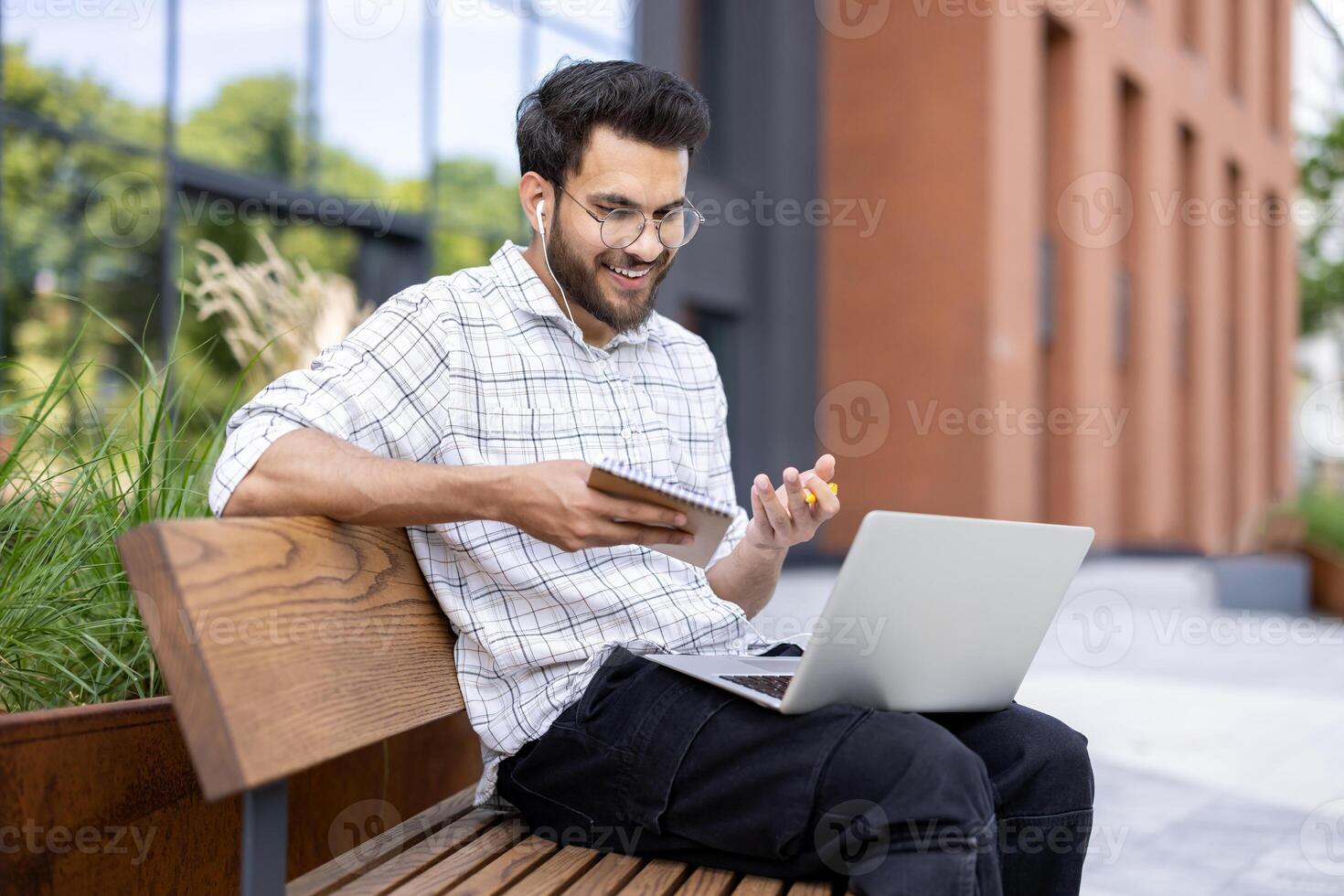 Indian smiling man sitting outside on the street on a bench, holding a notebook with a pen in his hands, talking on a call through a laptop. photo