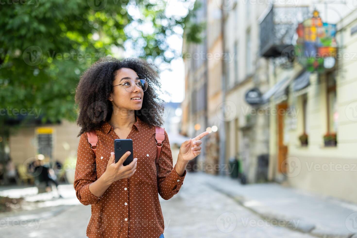 Woman exploring city street holding phone, pointing at building, wearing glasses and backpack. Concept of adventure, tourism, technology, and exploration. Outdoors urban environment on sunny day. photo