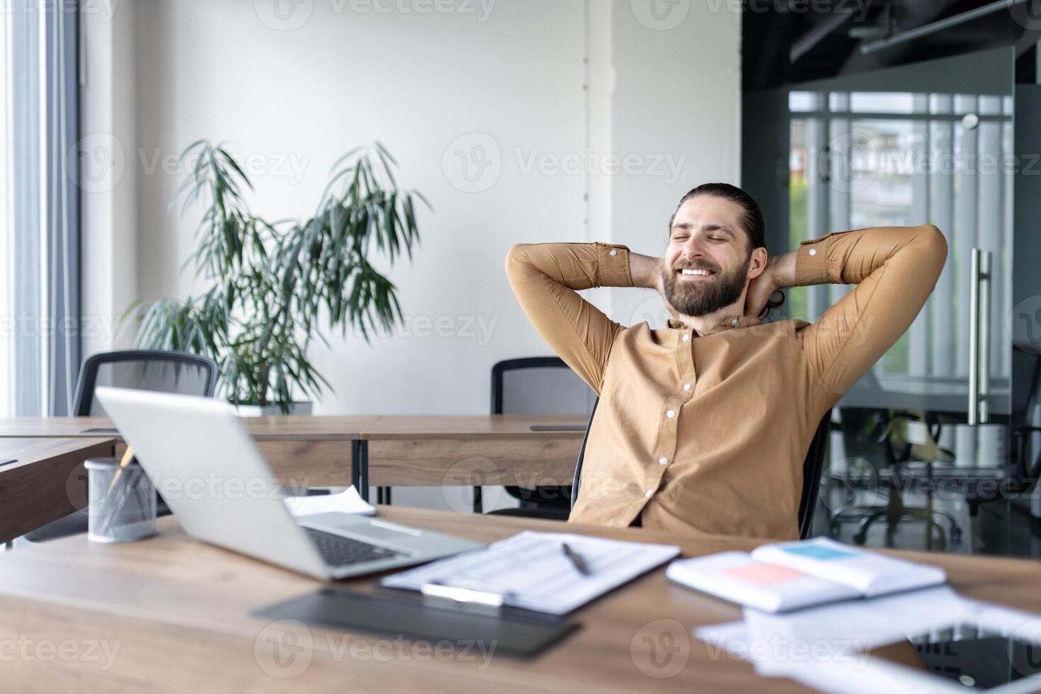 Businessman relaxing in office with hands behind head. Modern workspace features laptop, documents, indoor plants. Scene conveys workplace comfort, stress relief, professional satisfaction. photo