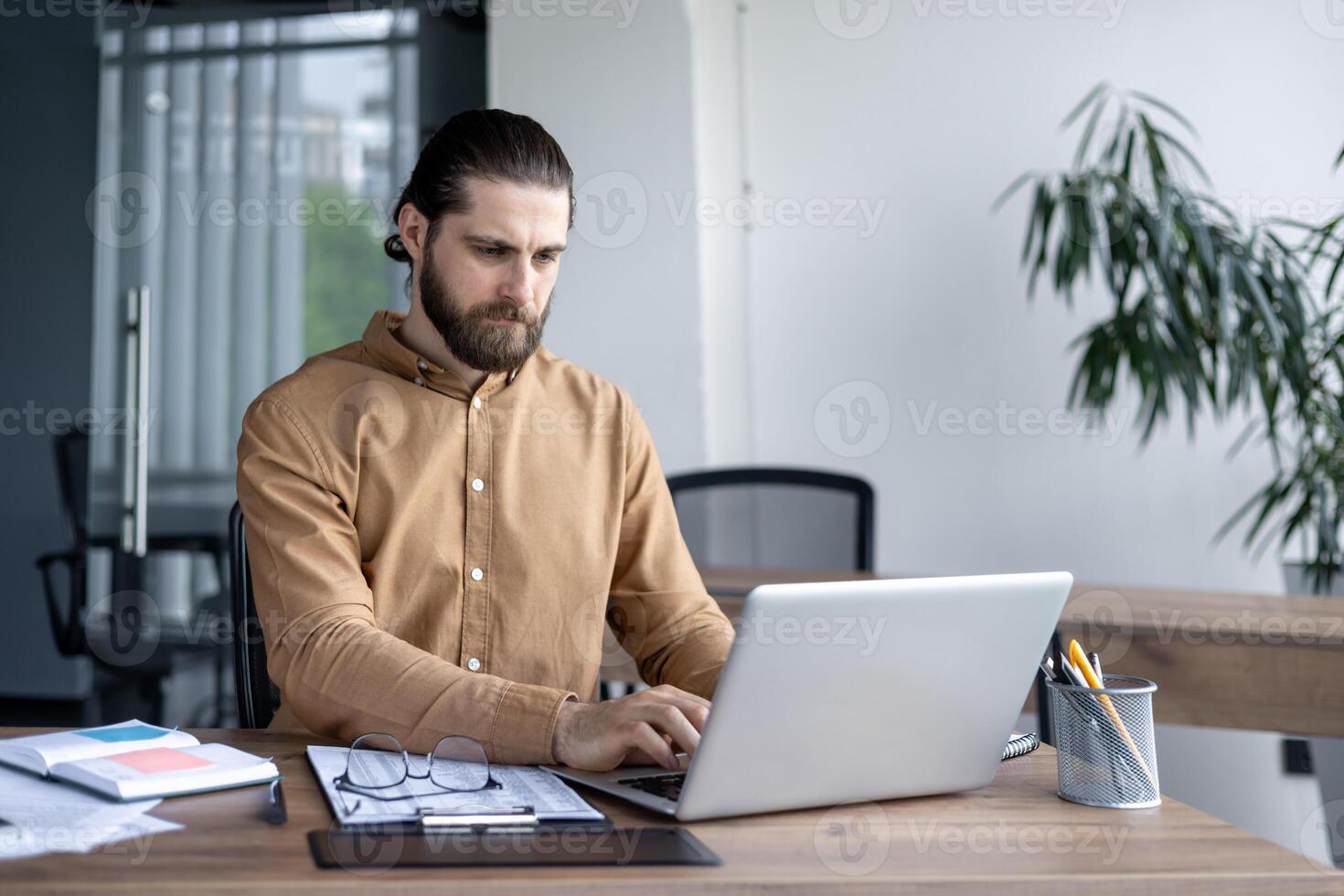 Professional male office worker using laptop at desk. Man concentrates on tasks surrounded by documents and office supplies. Modern workplace setting with natural light and plants photo