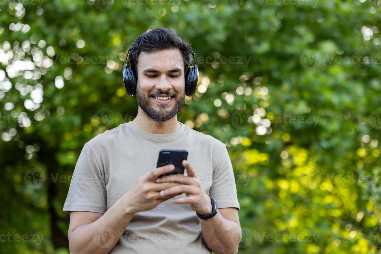 Young man wearing headphones smiling at smartphone in park. Relaxed outdoor setting captures enjoyment, technology, leisure, and connection with nature on sunny day. photo