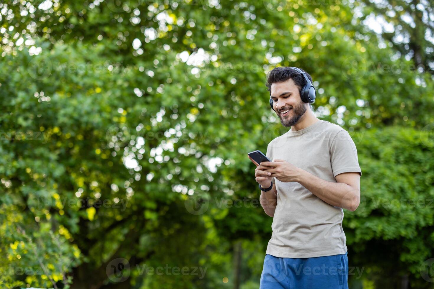 Young man in park using smartphone while listening to music with headphones. He looks relaxed and happy, enjoying moment outdoors in nature. Captures technology, leisure, and peacefulness photo