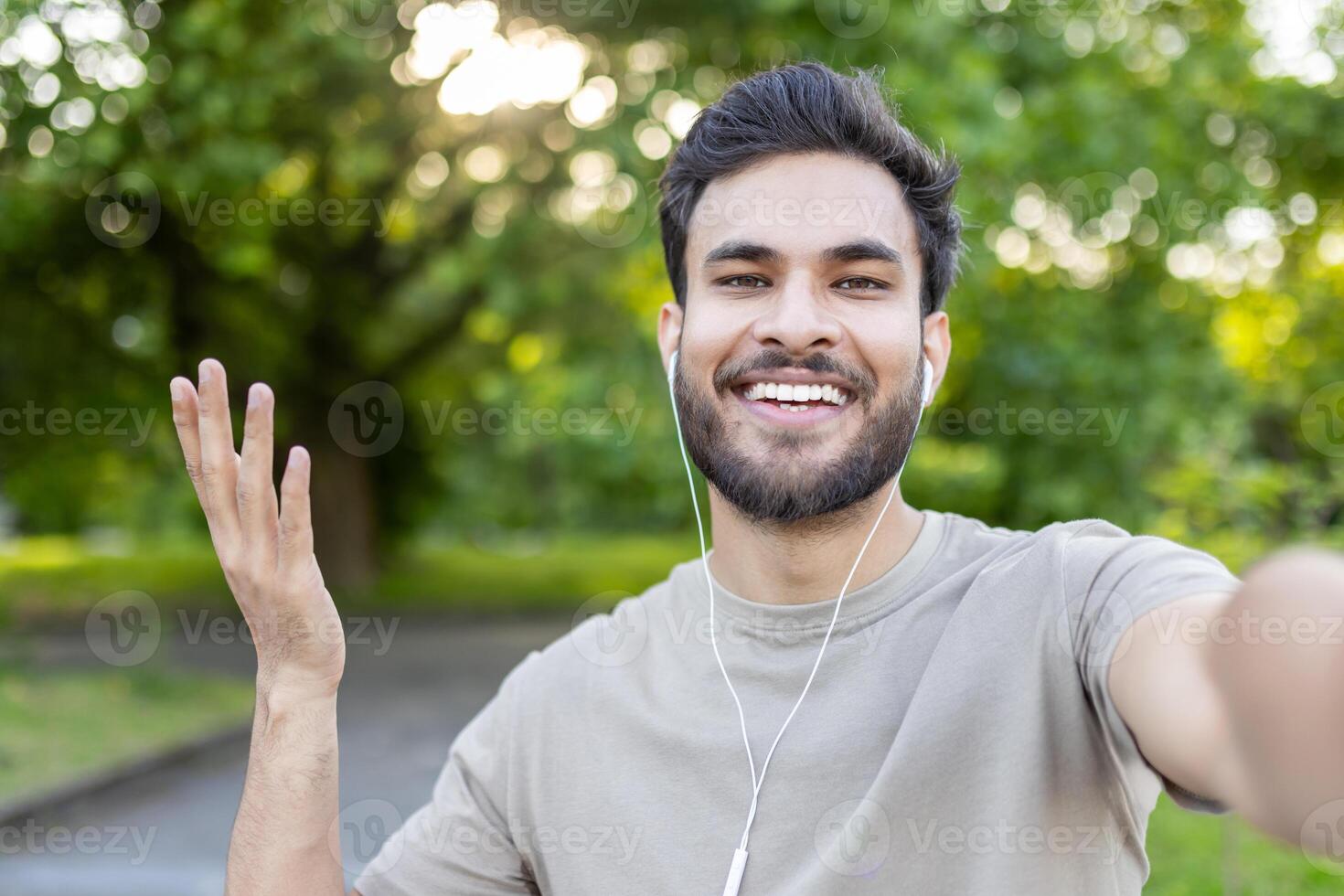 Young man smiling while taking selfie in park with smartphone and earphones. Captures happiness, connection, and relaxed outdoor atmosphere during leisure time. photo