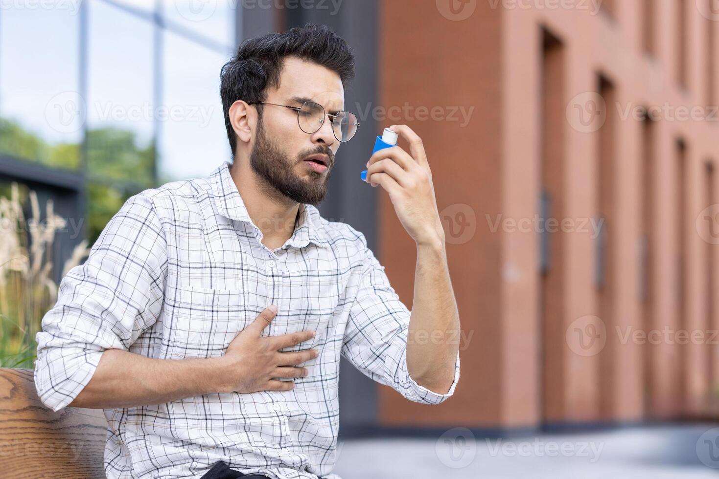Young man seated outdoors using asthma inhaler for respiratory relief. Wearing glasses and plaid shirt, holding inhaler and chest. Outdoor urban environment with greenery and buildings in background. photo