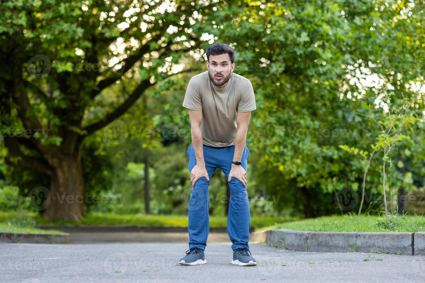Young sportsman resting after an intense exercise session in park. Dressed in casual sportswear, he leans forward catching breath, surrounded by serene green environment. photo