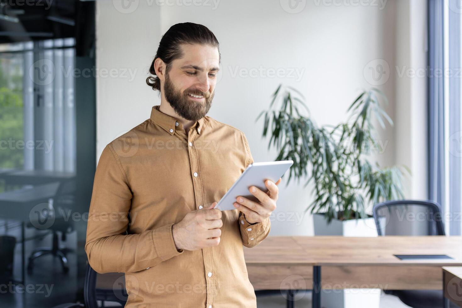 Young man with beard holding tablet in office setting. Smiling, engaged with digital device, reflecting modern business communication style. Office workspace hints collaboration and technology-driven photo