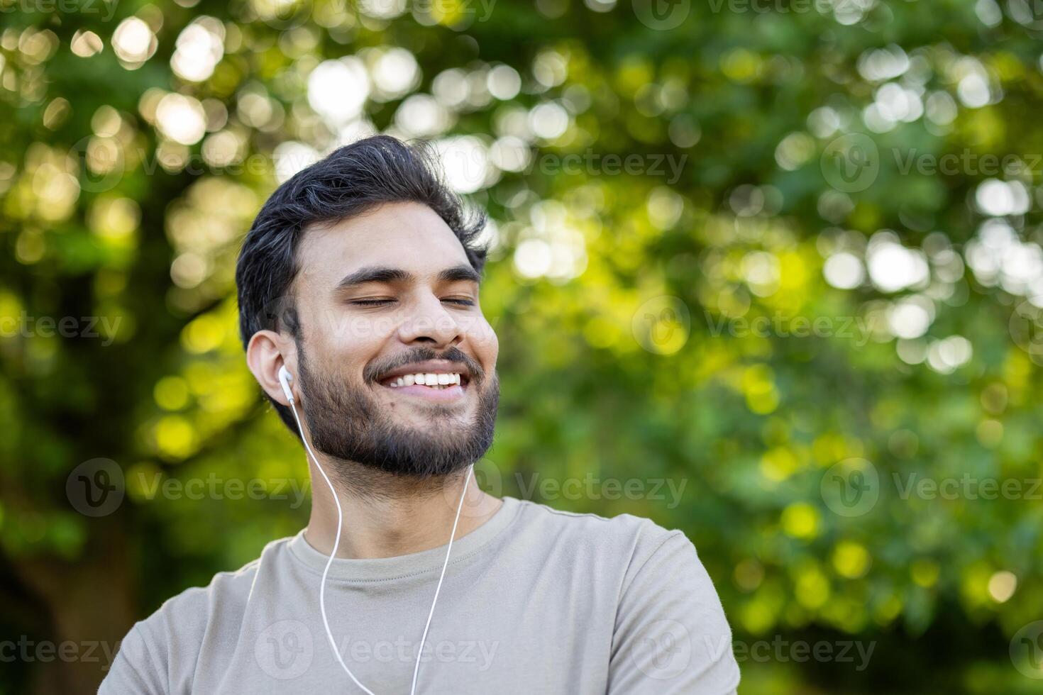 Young man smiling with closed eyes wearing headphones in green park. Enjoying music with relaxed and content expression. Sunlit background offers peaceful, serene atmosphere promoting mindfulness photo