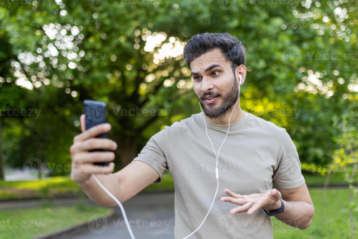 Young man in casual attire having energetic call in park setting. Holding smartphone, wearing earphones, expressing engaging emotions and gestures. Capturing essence of modern communication photo
