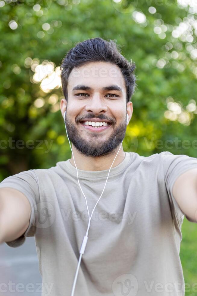 Cheerful young man smiling while taking selfie outdoors in park. He is wearing earbuds, enjoying nature and fresh air. Represents technology, happiness, and healthy lifestyle in natural setting. photo