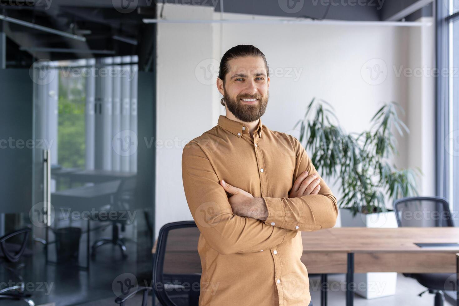 A cheerful, bearded businessman standing confidently with arms crossed in a contemporary office environment, exuding professionalism and positivity. photo