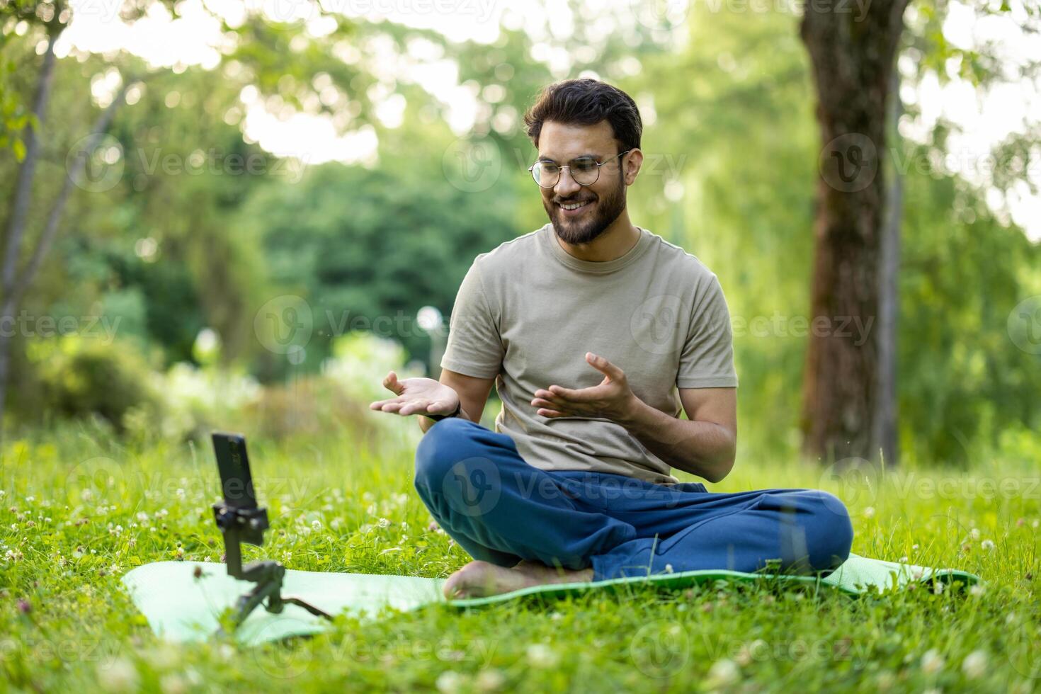 Smiling young man sits cross-legged on mat in park, conducting online yoga session with smartphone. Demonstrates mindfulness, relaxation techniques surrounded by greenery. Engaging with audience photo