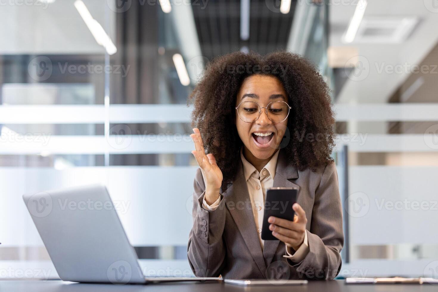 Young African American businesswoman in office using phone, expressing excitement, and gesturing. Professional attire, engaging with technology at workplace. Happy moment captured during work. photo