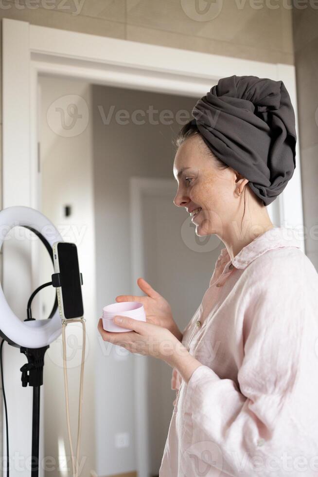 Blogger smiling and showing cosmetic cream jar in bathroom in front of phone photo