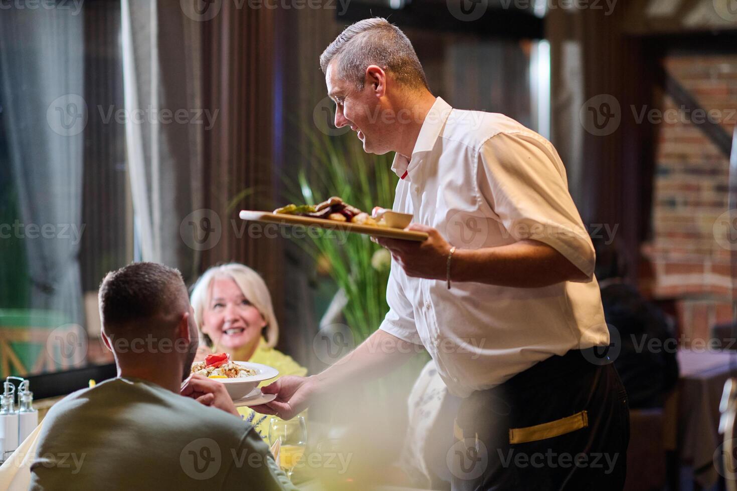 A courteous waiter provides impeccable service to an elderly family and their friends, creating a convivial atmosphere as they gather for a shared lunch, indulging in the diverse and delectable photo