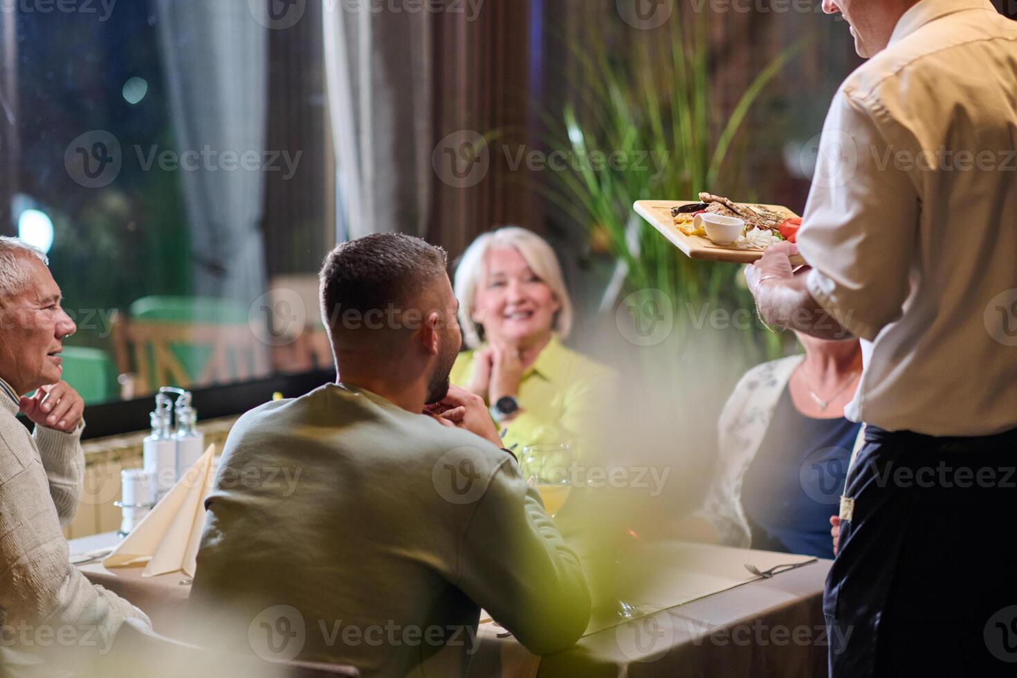A courteous waiter provides impeccable service to an elderly family and their friends, creating a convivial atmosphere as they gather for a shared lunch, indulging in the diverse and delectable photo