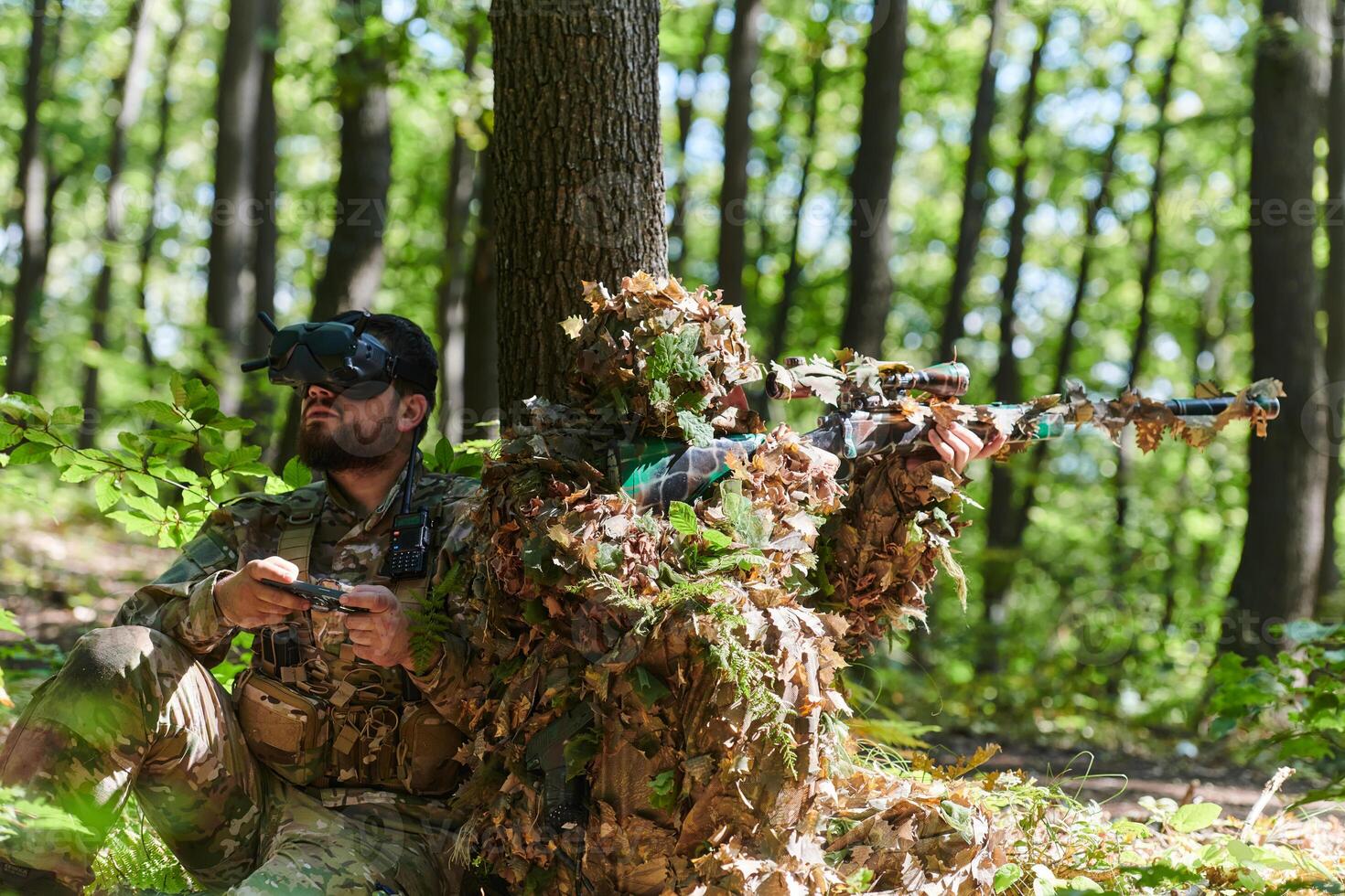 A skilled sniper and a soldier operating a drone with VR goggles strategize and observe the military action while concealed in the forest photo