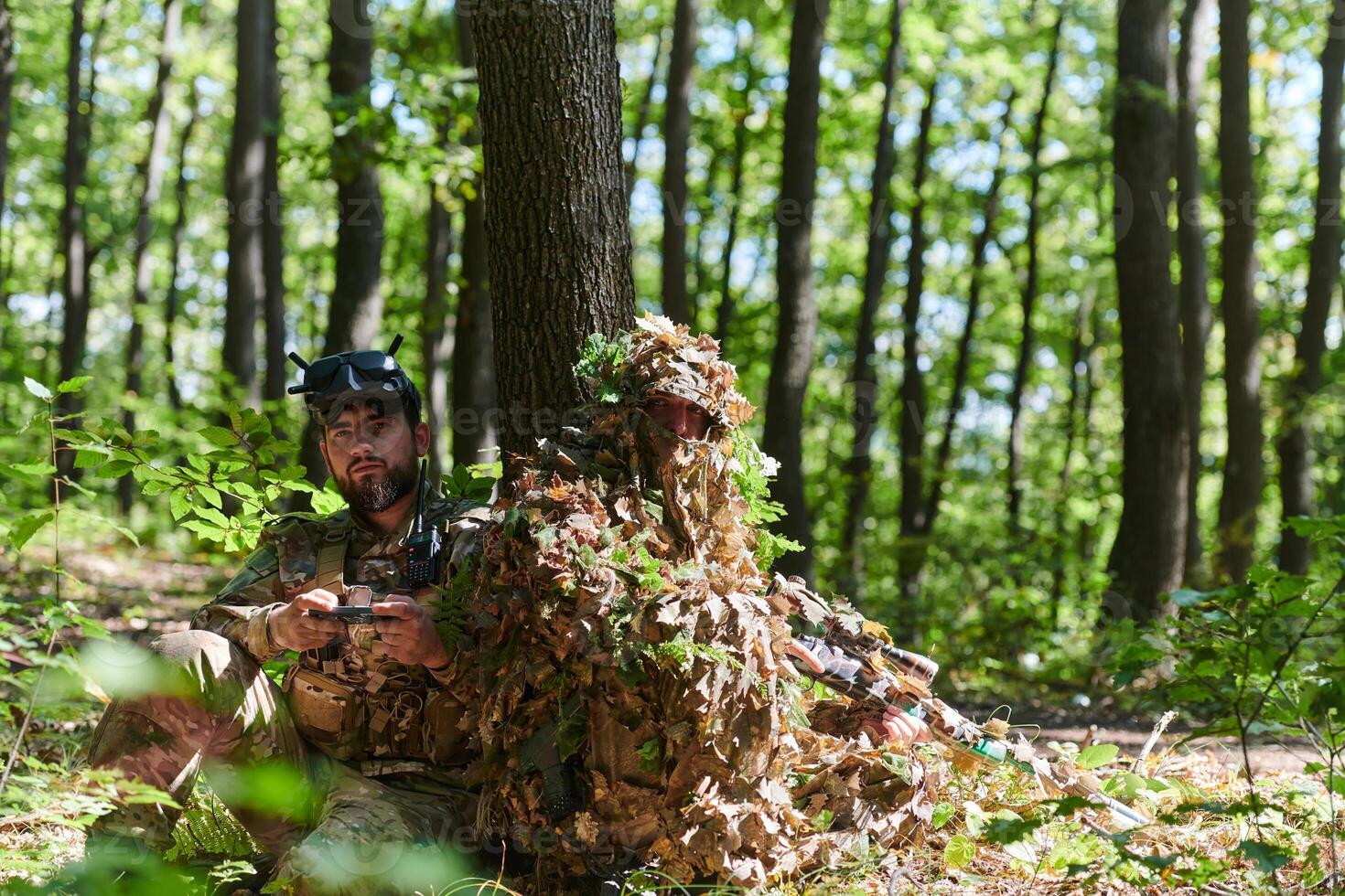 A skilled sniper and a soldier operating a drone with VR goggles strategize and observe the military action while concealed in the forest photo
