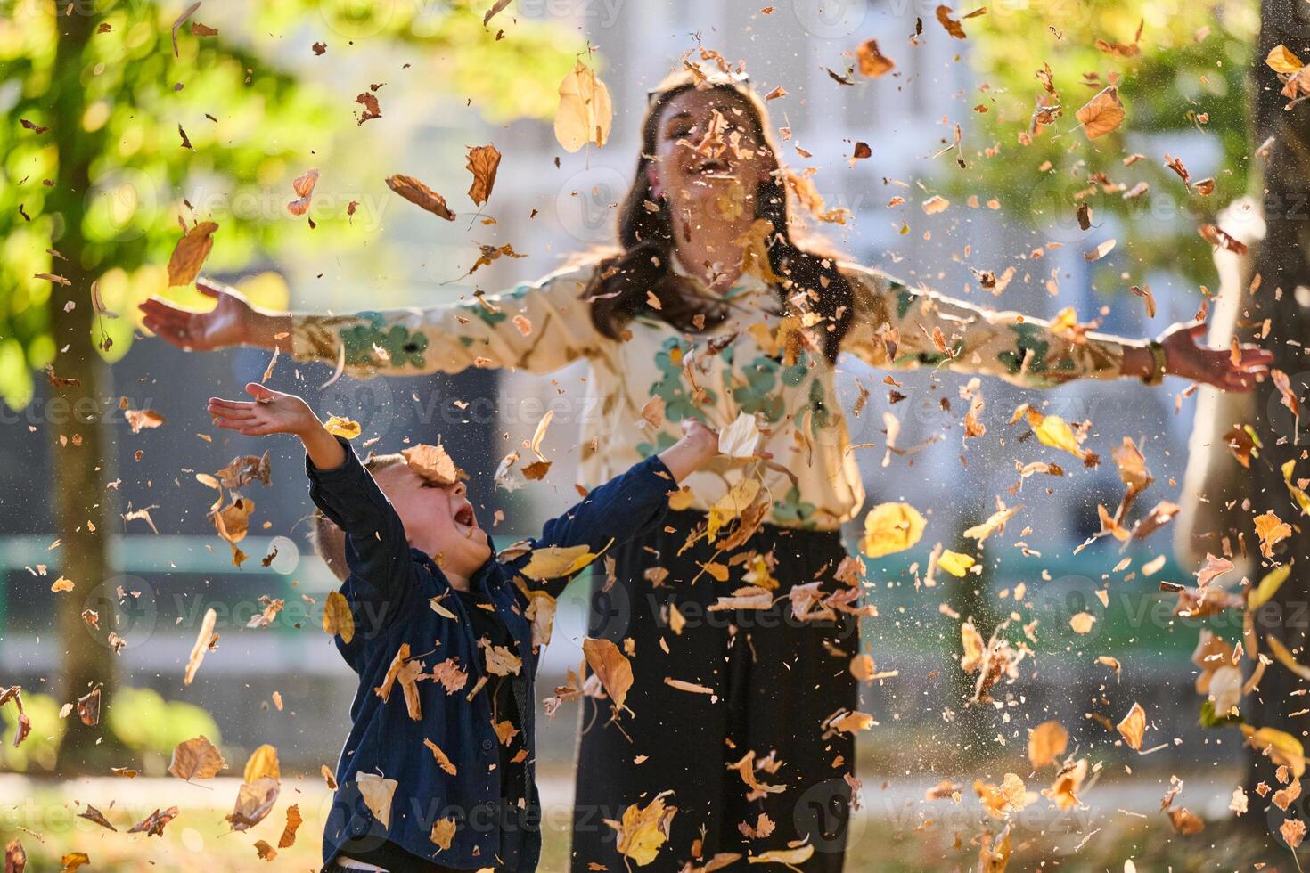 A modern woman joyfully plays with her son in the park, tossing leaves on a beautiful autumn day, capturing the essence of family life and the warmth of mother-son bonding in the midst of the fall photo