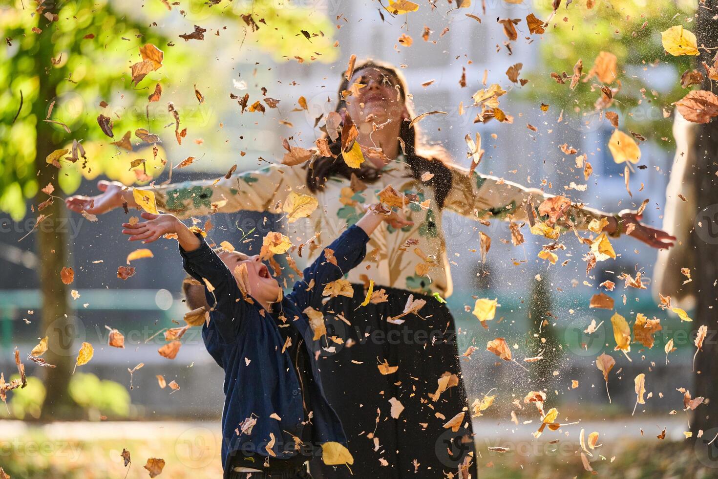 A modern woman joyfully plays with her son in the park, tossing leaves on a beautiful autumn day, capturing the essence of family life and the warmth of mother-son bonding in the midst of the fall photo