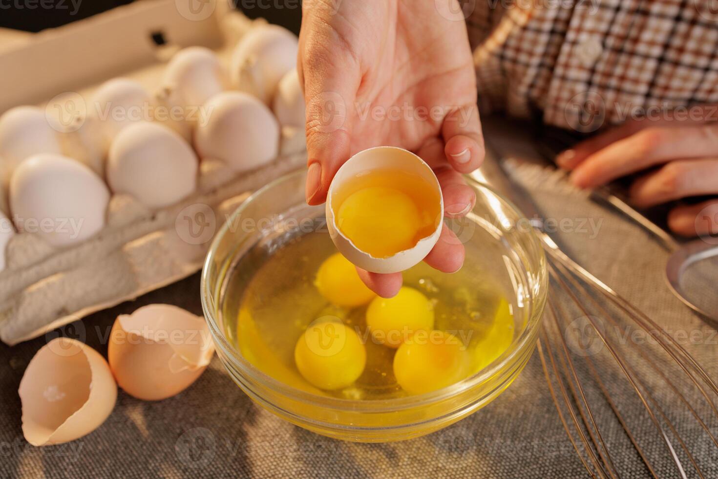 A person cracking an egg into a glass bowl, with several eggs in an egg carton in the background photo