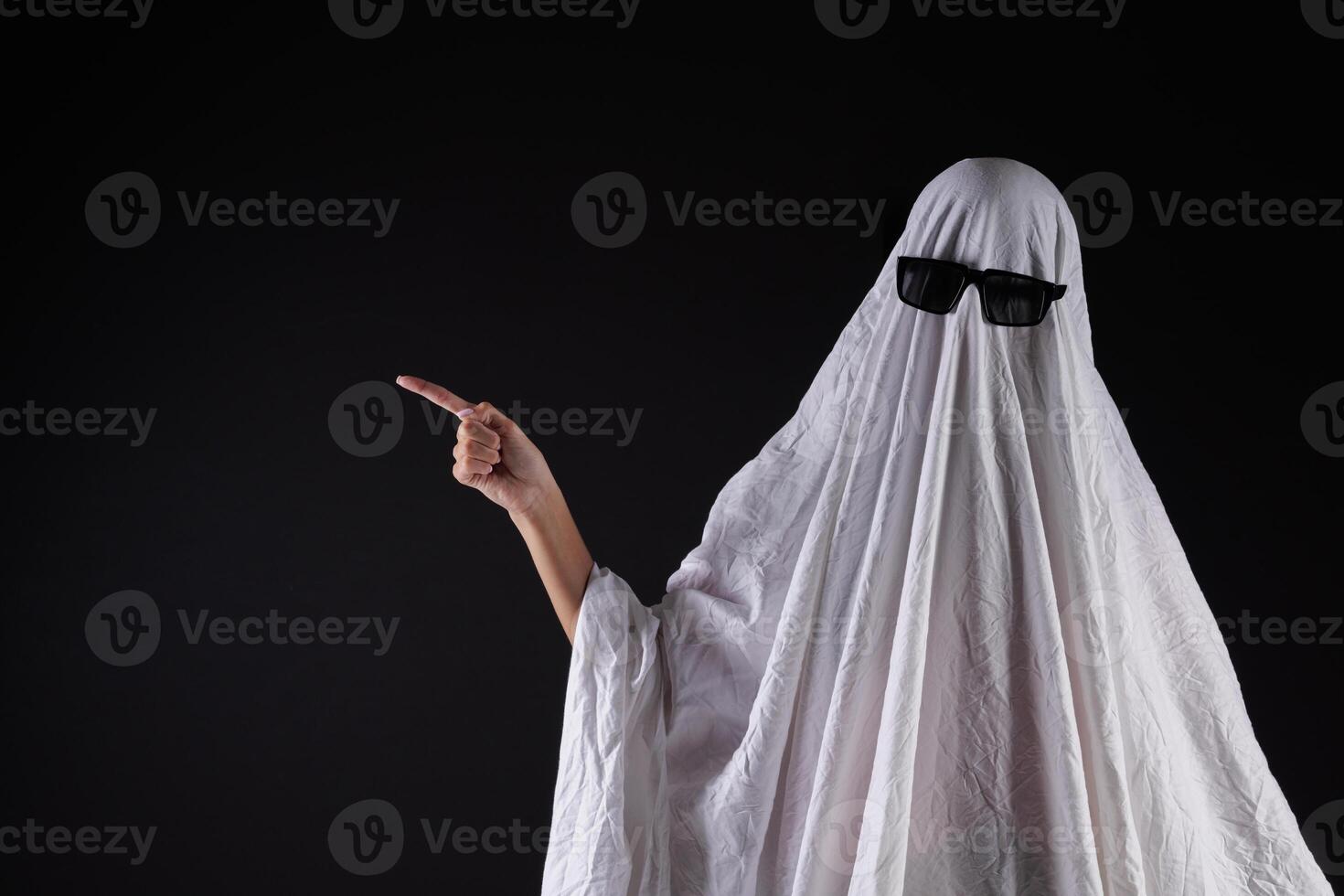 Person in costume of ghost pointing at something on dark background with space for tex photo