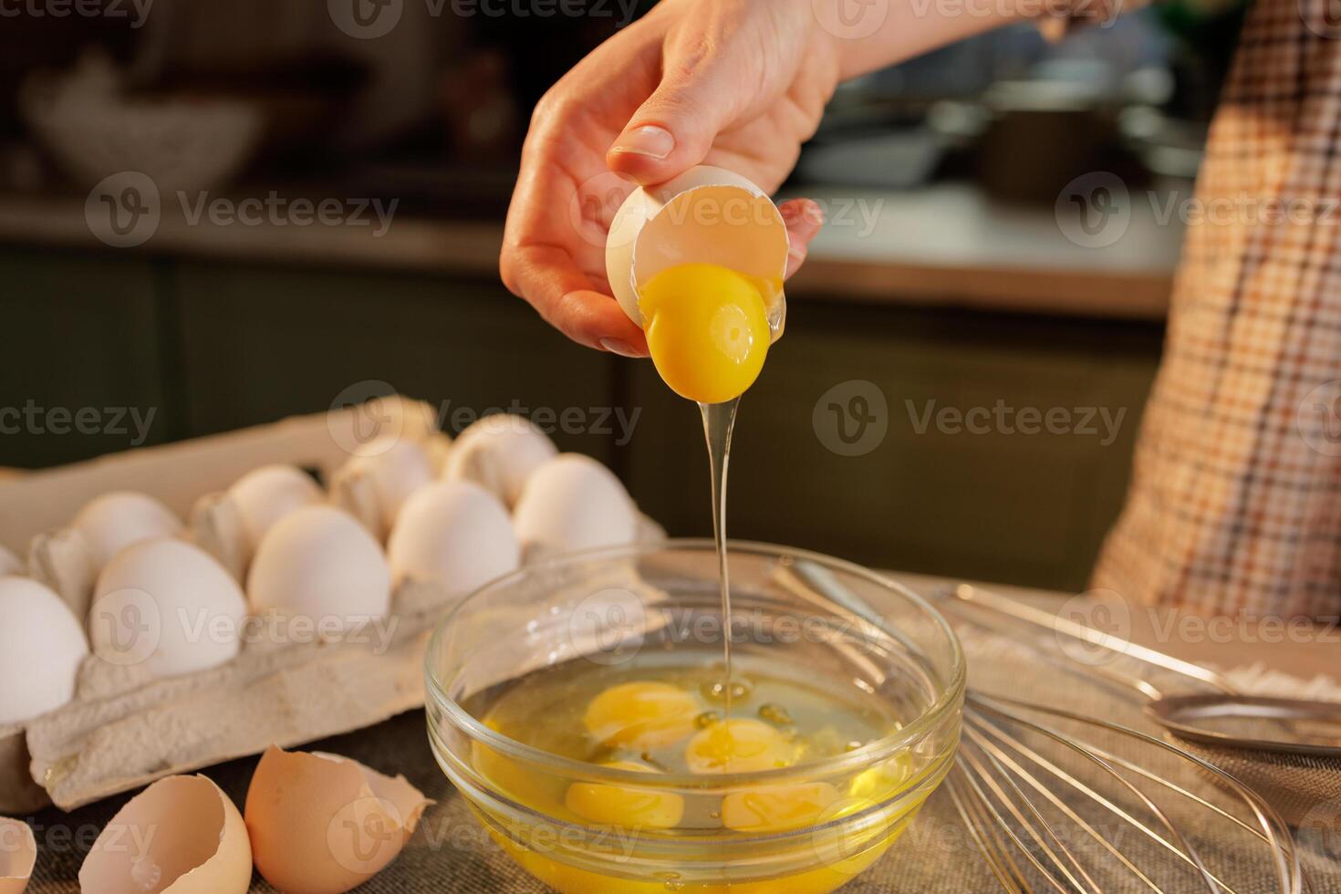A person cracking an egg into a glass bowl, with several eggs in an egg carton in the background photo