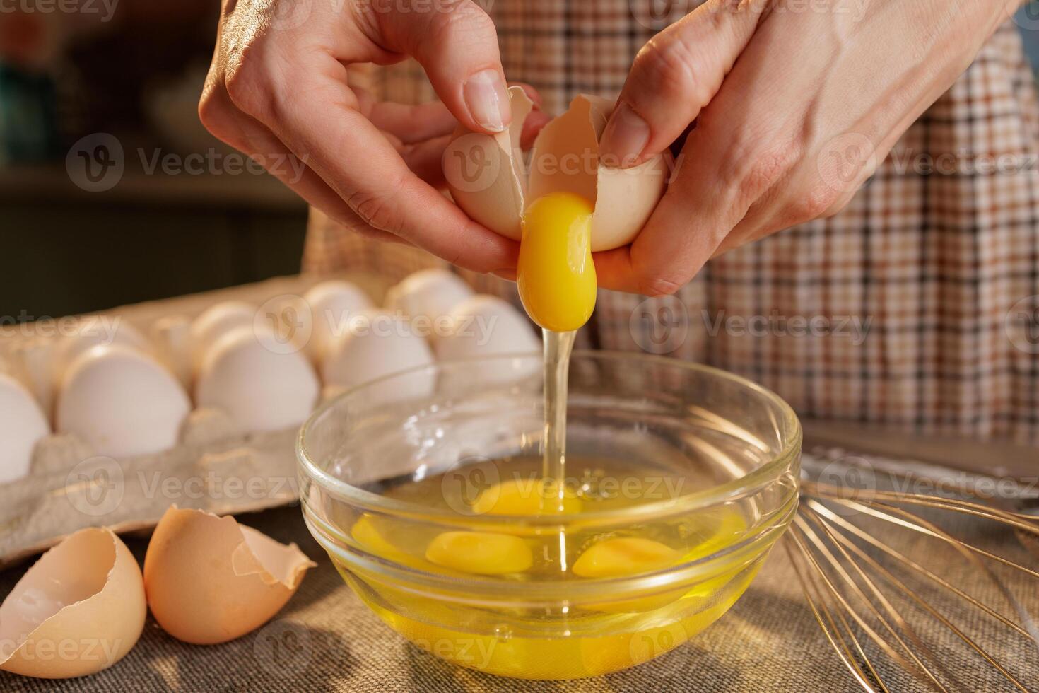 A person cracking an egg into a glass bowl, with several eggs in an egg carton in the background photo